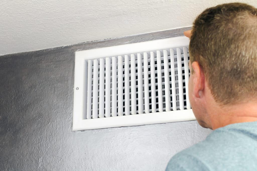 technician looking inside an upper wall white grid air duct on a silver wall near a white ceiling while performing air duct repair